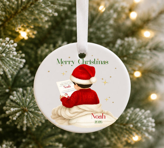 A personalised round ceramic Christmas ornament hanging from a white ribbon, featuring an illustration of a child in a red sweater and Santa hat reading a book, with the text 'Merry Christmas' and 'Noah 2025', set against a blurred Christmas tree background with warm lights.