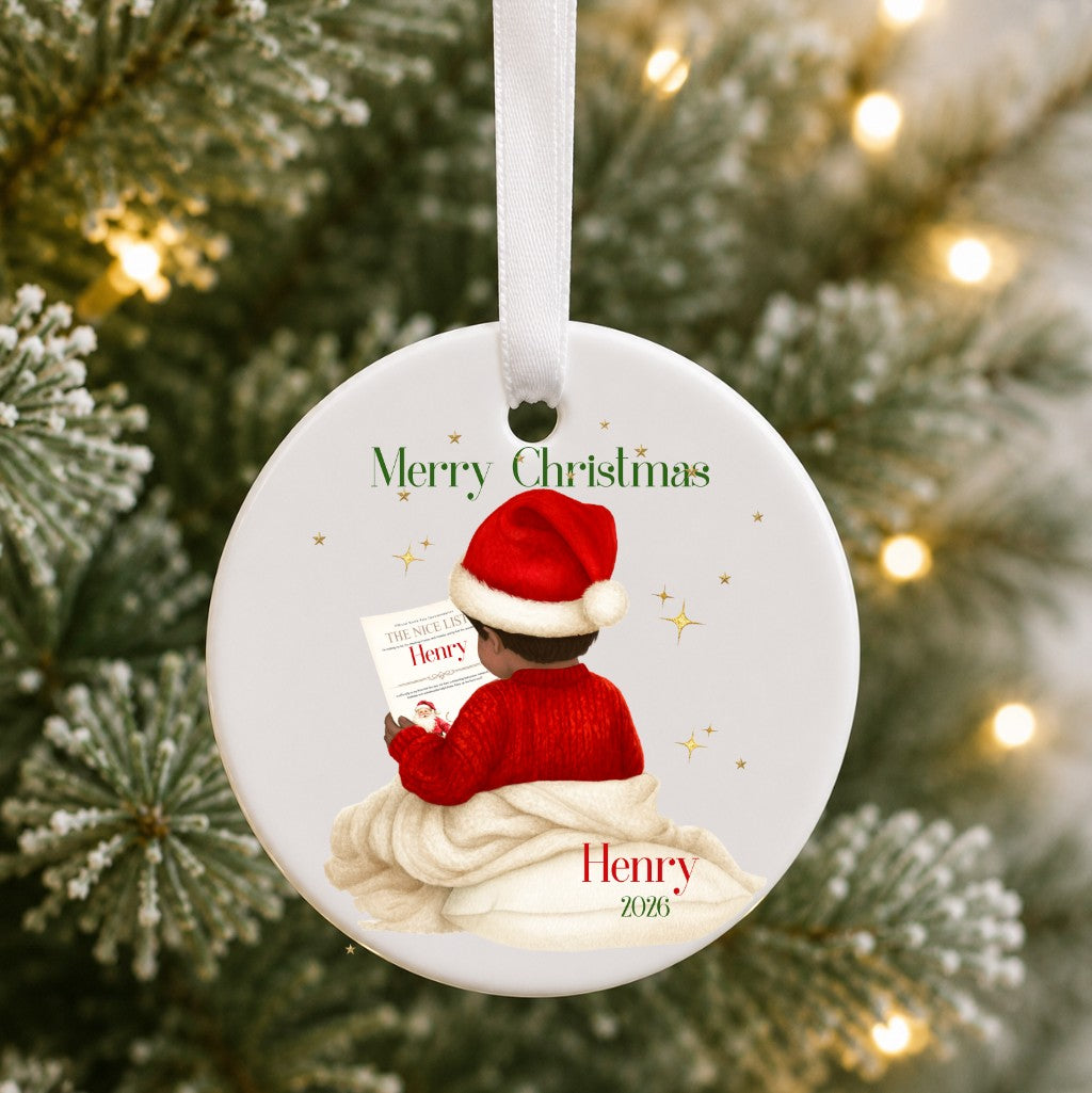 A round white ceramic Christmas ornament hanging from a white ribbon, featuring an illustration of a child in a red Santa hat and red sweater sitting on a cream blanket and reading a letter. The ornament is personalized with the text 'Merry Christmas', 'Henry', and '2025'.