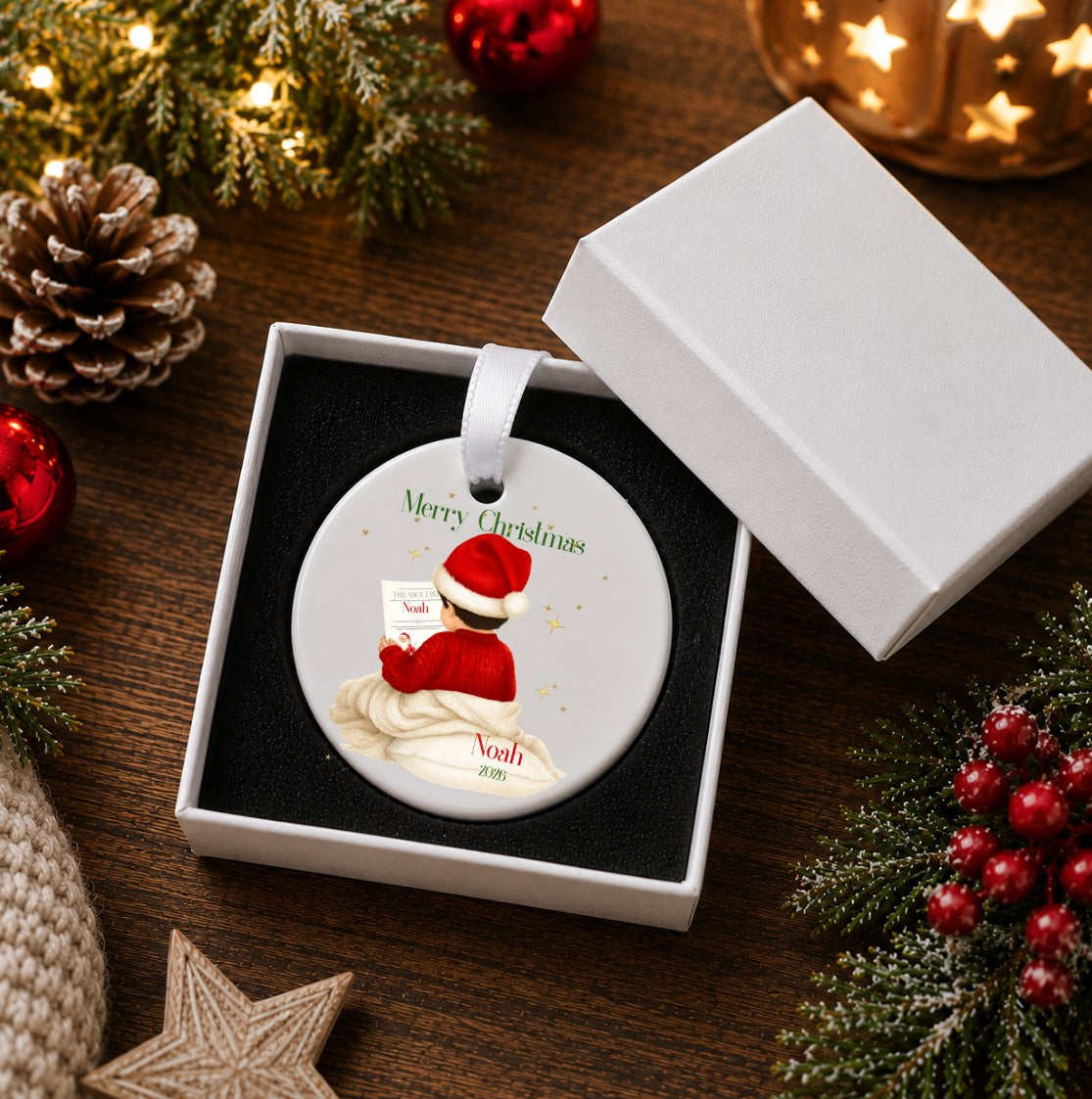 A round personalised Christmas ornament displayed in an open white gift box on a dark wooden surface, surrounded by holiday decorations including pine branches, red ornaments, a pinecone, and a star. The ornament features an illustration of a person in a Santa hat reading a letter, with the text 'Merry Christmas' and a custom name.