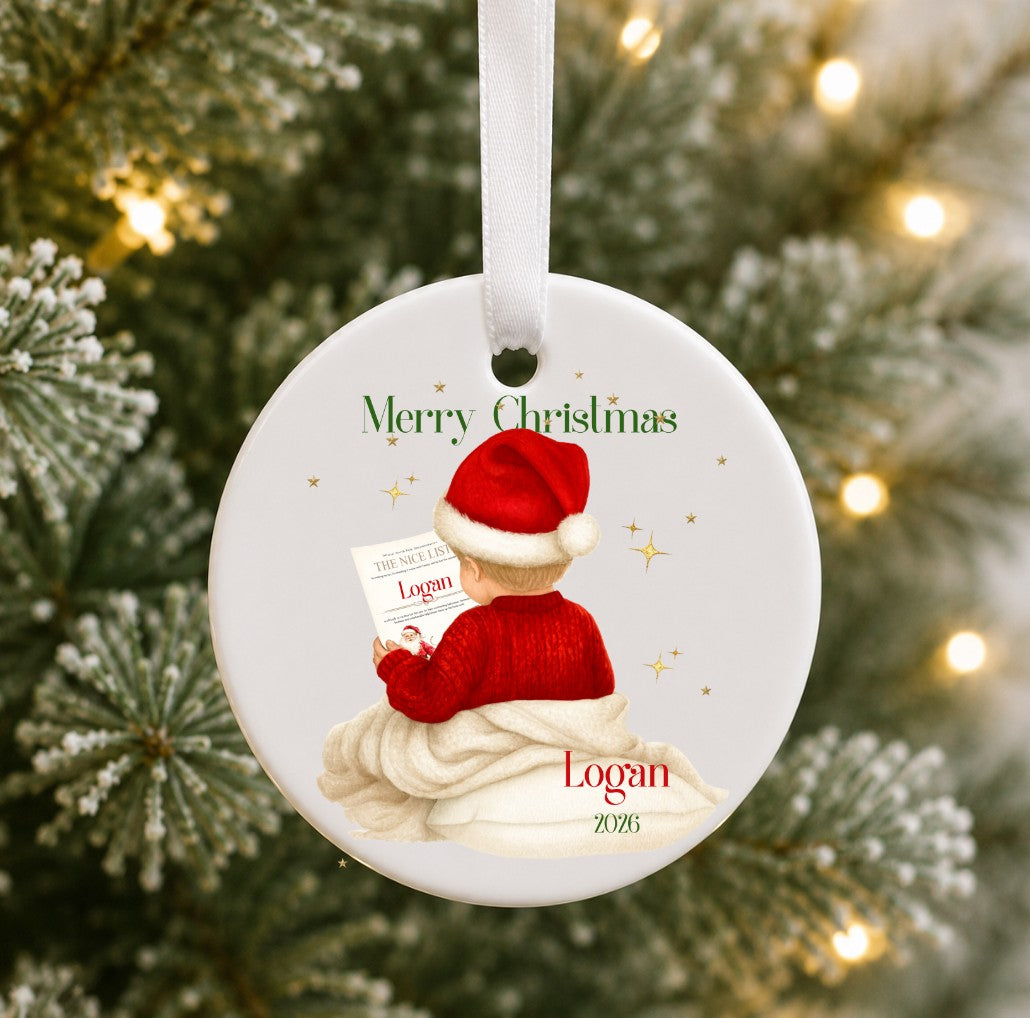A personalised round ceramic Christmas ornament hanging from a white ribbon, featuring an illustration of a baby in a Santa hat reading a book, with the text 'Merry Christmas' and the name 'Logan' on a blurred Christmas tree background with warm lights.