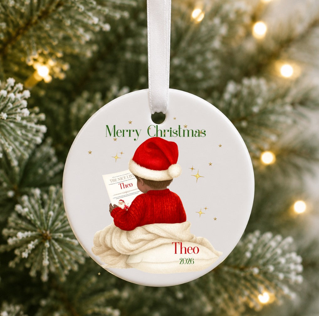 A personalised round ceramic Christmas ornament hanging from a white ribbon, featuring an illustration of a child in a red sweater and Santa hat reading a letter, with the text 'Merry Christmas' and 'Theo 2025'. The ornament is photographed against a blurred Christmas tree background with warm lights.