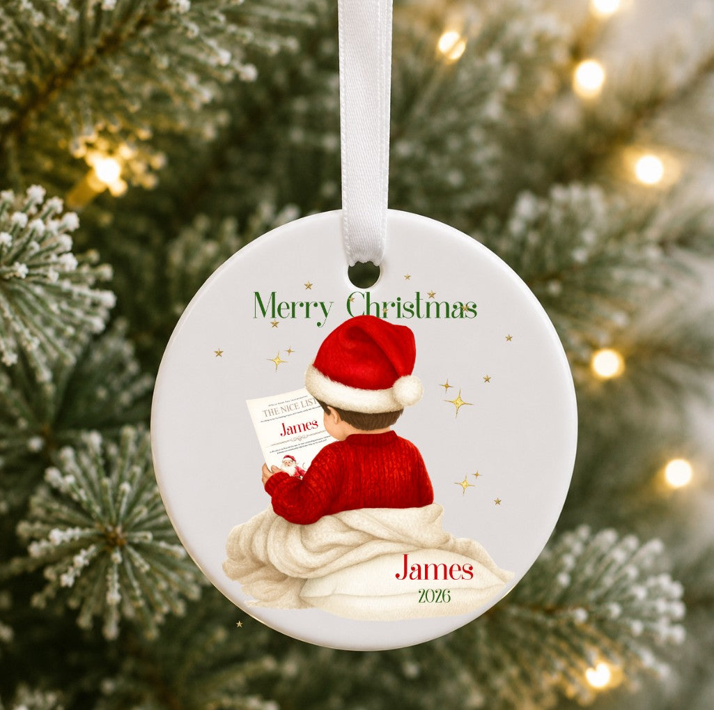 A personalised round ceramic Christmas ornament hanging from a white ribbon, featuring an illustration of a child in a red Santa hat and red sweater sitting in a white blanket and reading a letter addressed 'James'. The ornament is personalized with the name 'James' and the year '2025'.