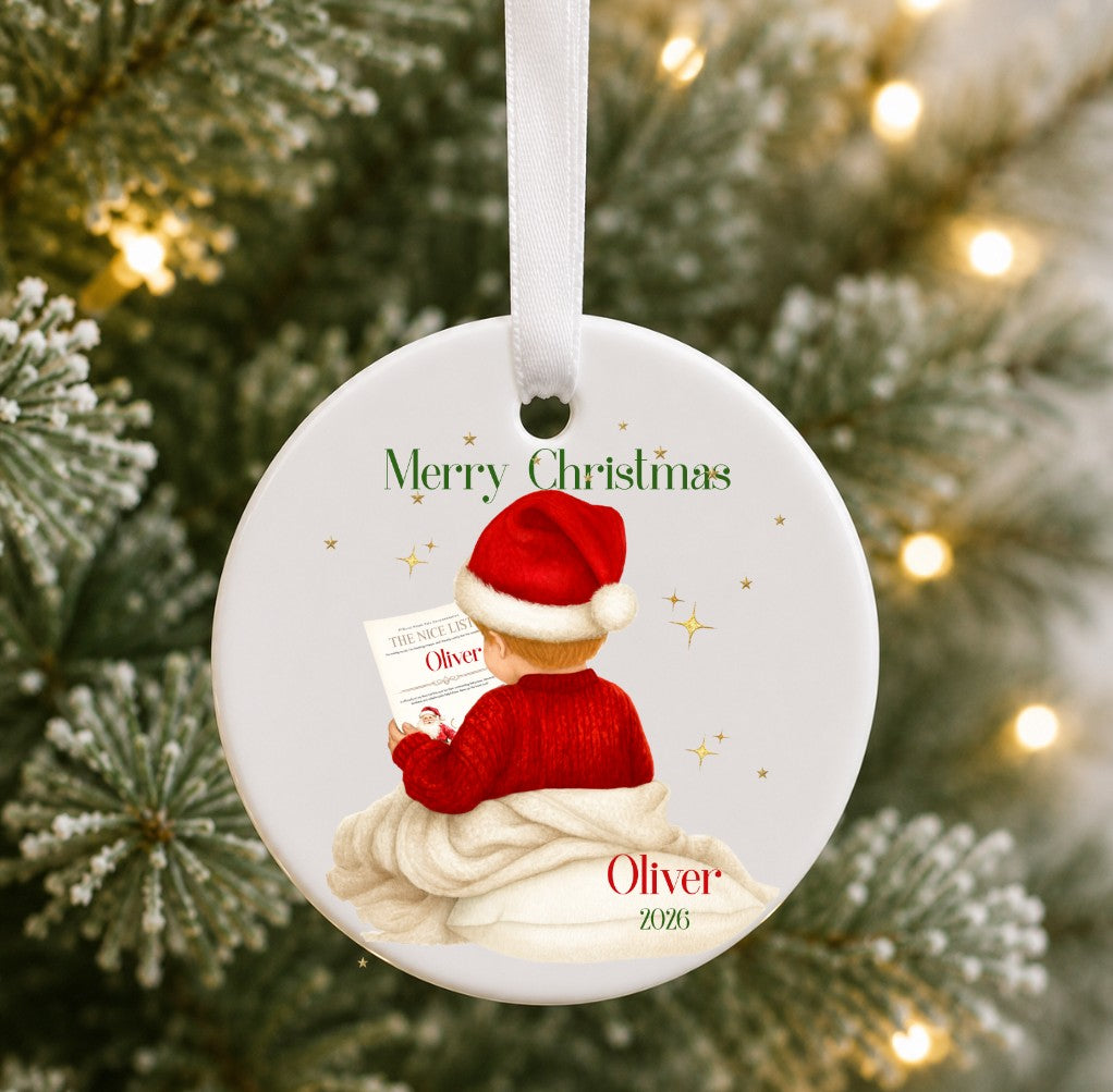 A personalised round white Christmas ornament hanging on a decorated Christmas tree. The ornament features an illustration of a child wearing a Santa hat and red sweater, sitting on a white blanket and reading a letter.
