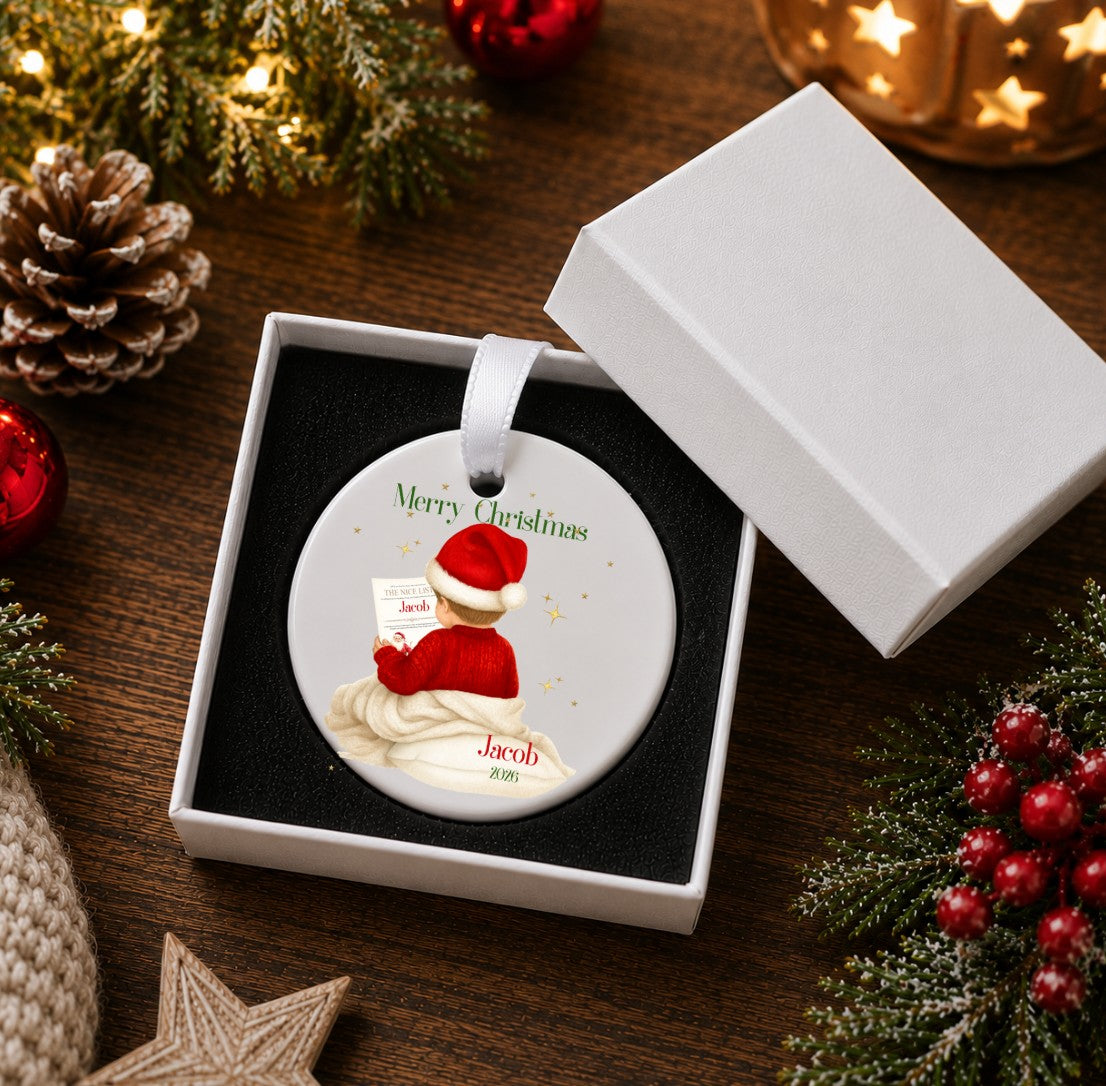 A personalised round Christmas ornament displayed in an open white gift box on a dark wooden table, surrounded by holiday decorations including pinecones, red berries, greenery, and warm string lights. The ornament features a Santa-clad figure with the text 'Merry Christmas' and the name 'Jacob'.