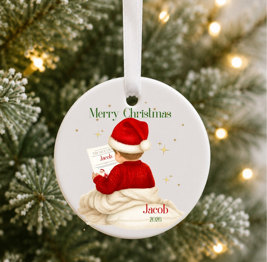 A personalised round Christmas ornament hanging on a blurred Christmas tree with warm lights. The ornament features an illustration of a child in a red Santa hat and red sweater sitting on a white blanket, reading a letter, with the text 'Merry Christmas' and the name 'Jacob 2025'.