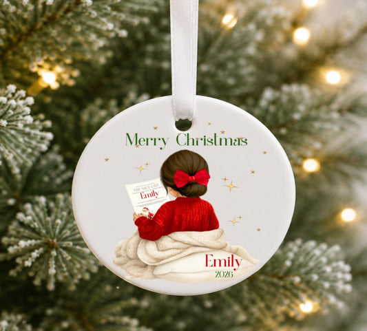 A personalised round Christmas ornament hanging on a decorated tree. The ornament features an illustration of a child with a red bow and red sweater sitting on a cream blanket, with the text 'Merry Christmas' and the name 'Emily 2026'.