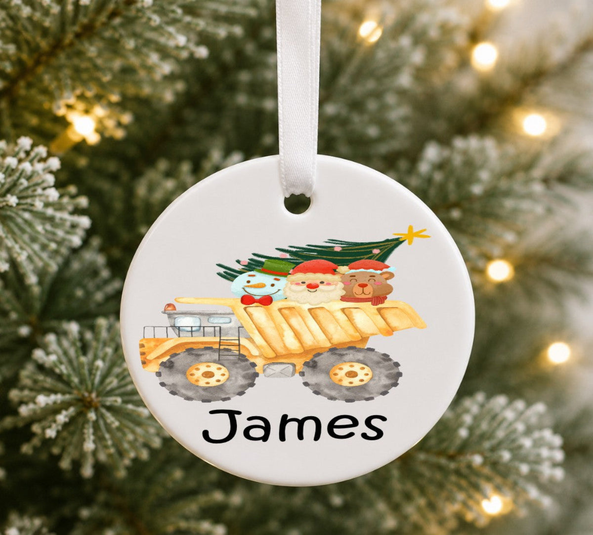 A round white ceramic Christmas ornament hanging from a white ribbon, printed with a cartoon snowman driving a yellow dump truck carrying a Christmas tree. The name 'James' is printed in black at the bottom.
