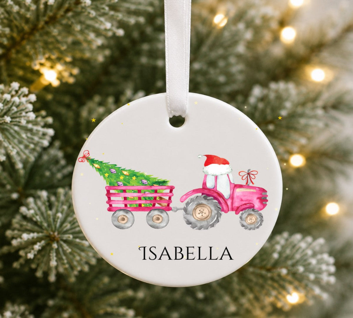 A personalised round white Christmas ornament hanging from a white ribbon, featuring a pink tractor and trailer with a green Christmas tree, the name 'ISABELLA' at the bottom, and a blurred festive tree background with warm lights.