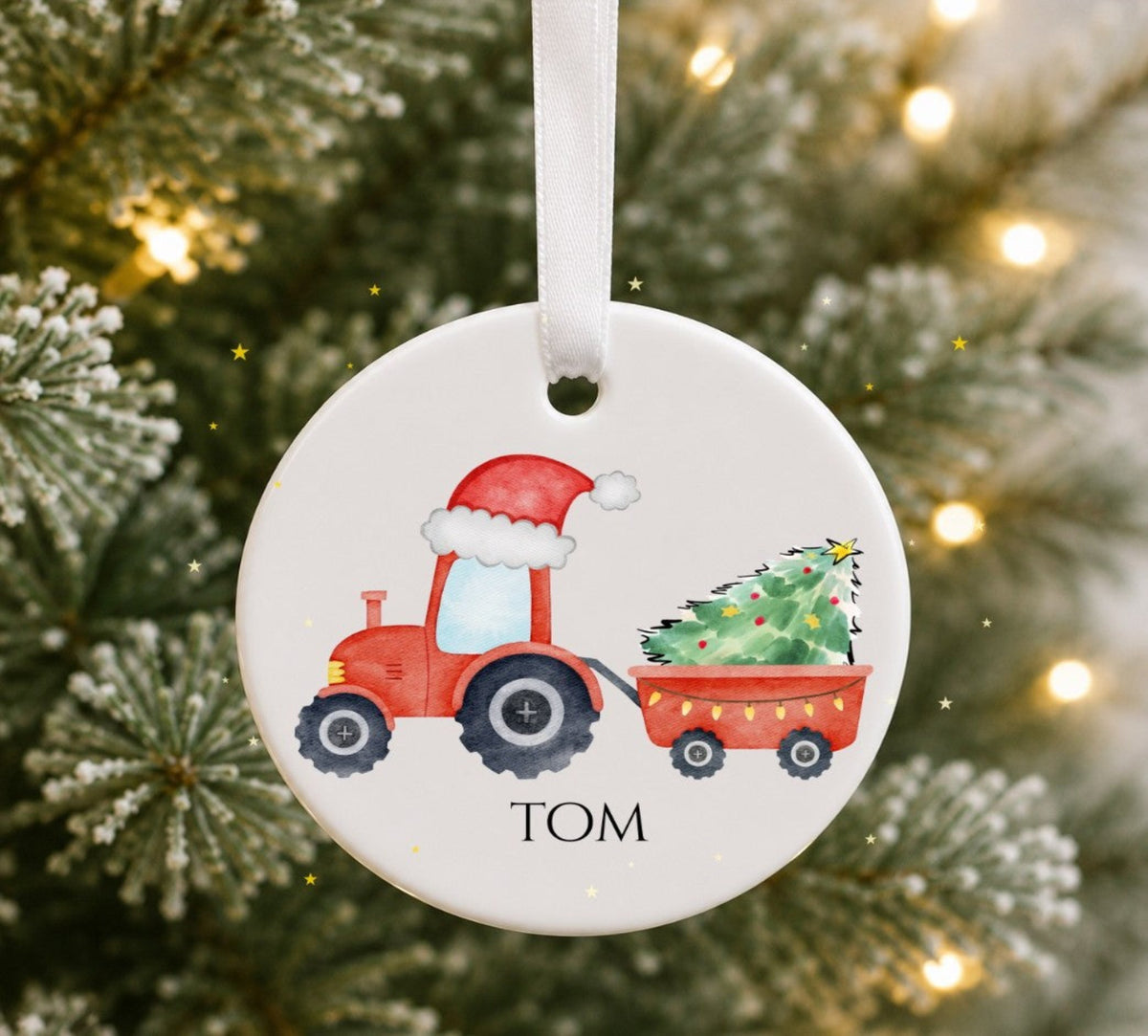 A round white ceramic Christmas ornament hanging from a white ribbon, featuring a red tractor with a Santa hat pulling a trailer with a Christmas tree, and the name 'TOM' printed below. The ornament is photographed in front of a blurred Christmas tree with warm lights.