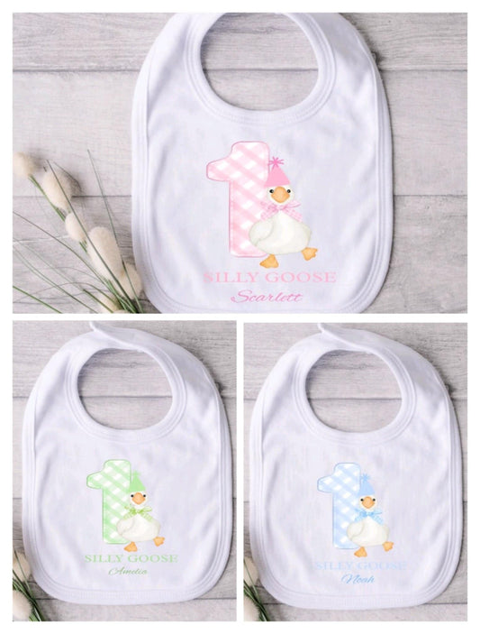 Three baby bibs with colourful goose designs and text on a wooden surface.