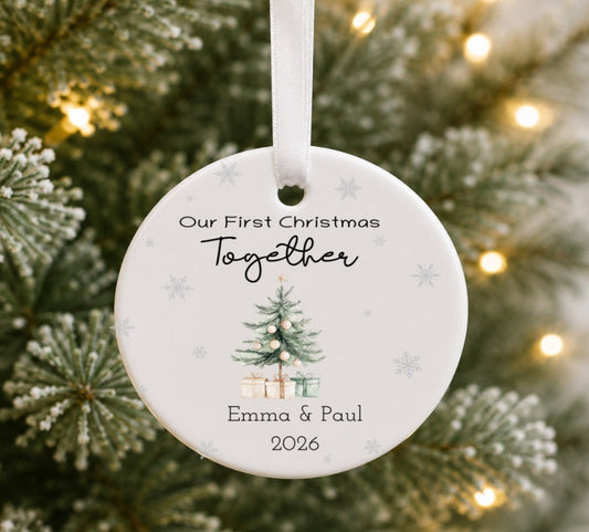 A round white ceramic Christmas ornament hanging from a white ribbon, printed with the personalised text 'Our First Christmas Together', a small illustrated Christmas tree with presents, and the names 'Emma & Paul' with '2026' below. The ornament is photographed hanging in front of a blurred Christmas tree with warm string lights.
