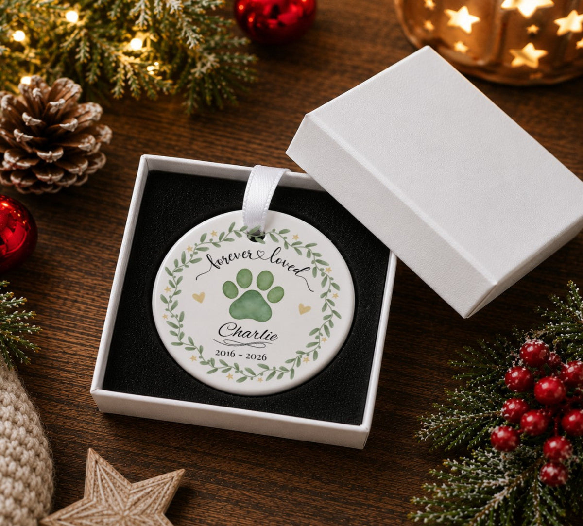 A round personalised Christmas ornament displayed in an open white gift box on a wooden surface, surrounded by holiday decorations including pine branches, red berries, a pinecone, and a glowing star lamp. The ornament features a white background with a printed illustration of a paw print.