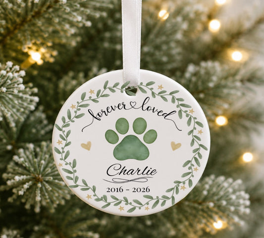 A round white pet memorial Christmas ornament hanging on a Christmas tree, featuring a green paw print, a leafy wreath border, and the text 'forever loved Charlie 2016 - 2026'.