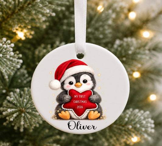 A round white ceramic Christmas ornament hanging from a white ribbon, featuring a cartoon penguin in a red Santa hat holding a red heart that reads 'MY FIRST CHRISTMAS 2026', with the name 'Oliver' printed below, set against a blurred Christmas tree background with warm lights.