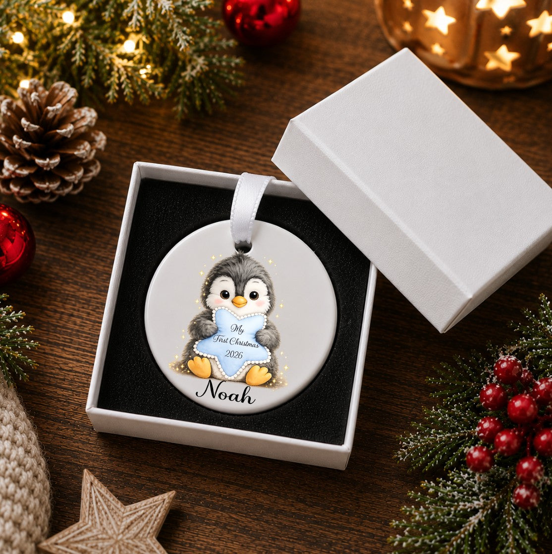 A personalised Christmas ornament featuring a cartoon penguin holding a banner that reads 'My First Christmas 2026' and the name 'Noah' below, presented in an open white gift box on a wooden table surrounded by holiday decorations.