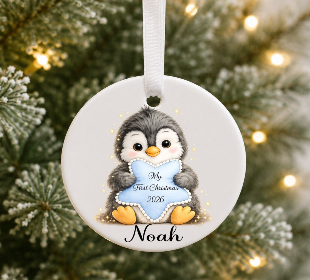 A personalised round ceramic Christmas ornament featuring a penguin holding a light blue heart that reads 'My First Christmas 2026' with the name 'Noah' below, hanging against a blurred Christmas tree background with warm lights.