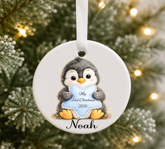 A personalised round ceramic Christmas ornament featuring a penguin holding a light blue heart that reads 'My First Christmas 2026' with the name 'Noah' below, hanging against a blurred Christmas tree background with warm lights.
