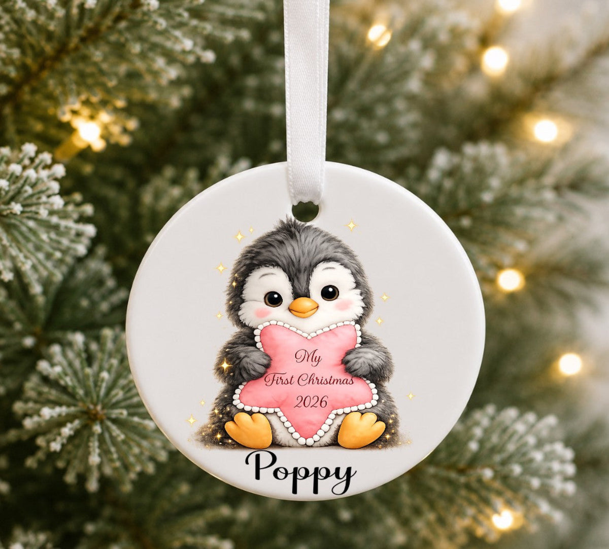 A personalised round ceramic Christmas ornament featuring a cute cartoon penguin holding a pink heart that reads 'My First Christmas 2026' with the name 'Poppy' below. The ornament hangs from a white ribbon against a blurred Christmas tree background with warm lights.