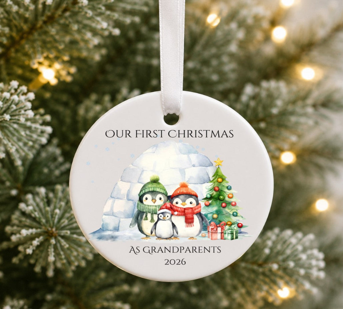 A round white ceramic Christmas ornament hanging from a white ribbon, printed with the text 'OUR FIRST CHRISTMAS' and 'AS GRANDPARENTS 2026' around an illustration of three penguins, an igloo, a Christmas tree, and wrapped gifts. The ornament is photographed in front of a blurred Christmas tree with warm lights.