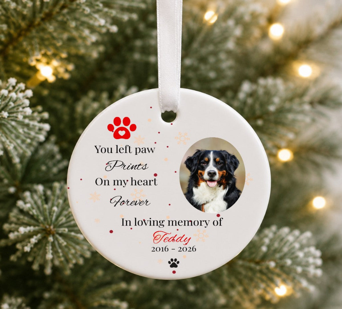 A personalised round white pet memorial Christmas ornament hanging on a Christmas tree with warm lights in the background. The ornament features a photo of a Bernese mountain dog, red paw print graphics, and printed text reading 'You left paw Prints On my heart Forever In loving memory of Teddy 2016 - 2026'.
