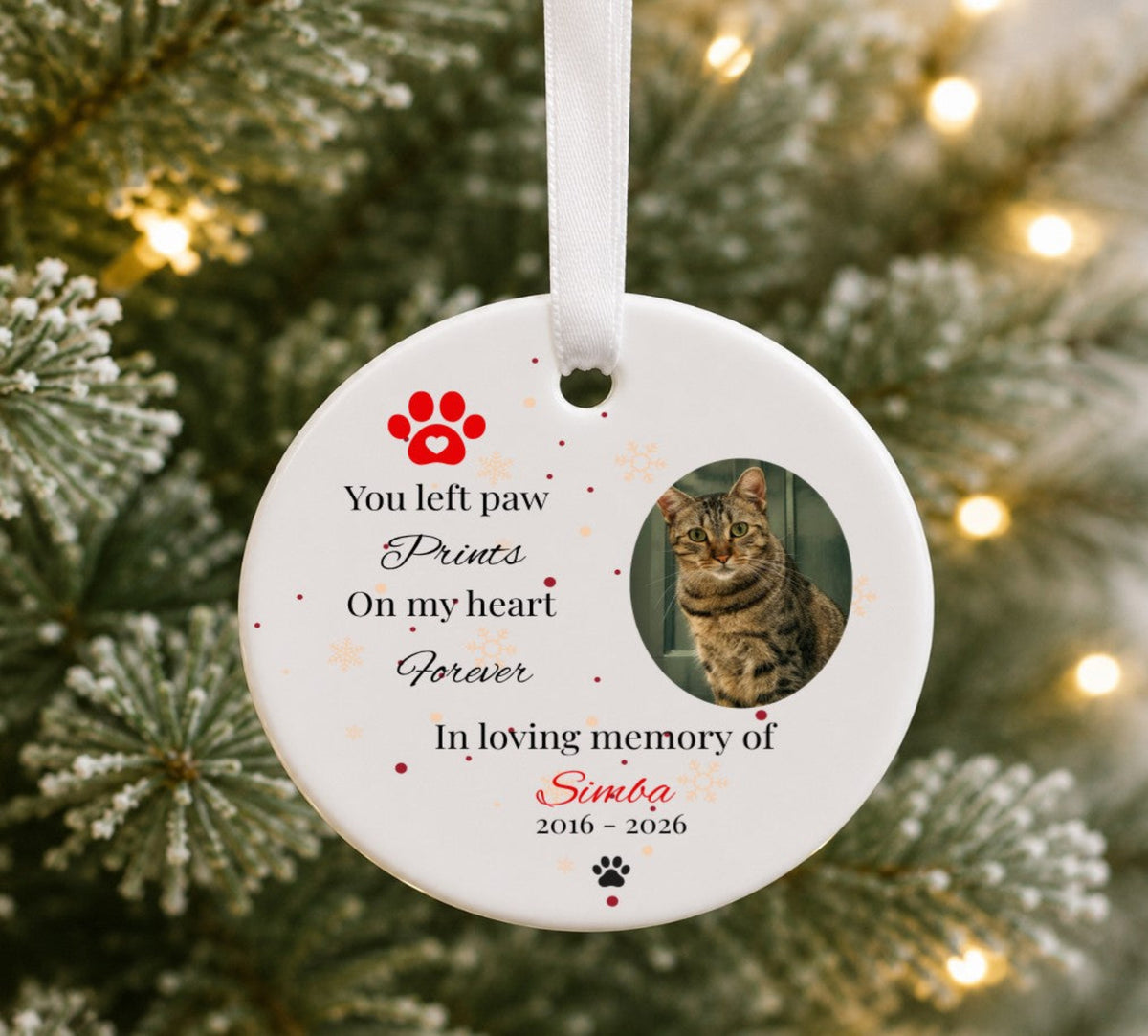 A personalised round white ceramic pet memorial Christmas ornament hanging on a blurred Christmas tree with warm lights. The ornament features a white ribbon, a red paw print, the text 'You left paw Prints On my heart Forever In loving memory of Simba 2016 - 2026', and a photo of a tabby cat.