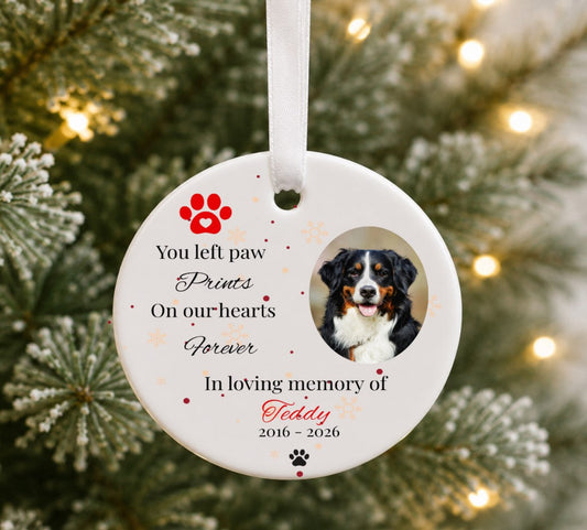 A personalised round white pet memorial Christmas ornament hanging on a Christmas tree with warm lights in the background. The ornament features a photo of a Bernese mountain dog, red paw print graphics, and printed text reading 'You left paw Prints On our hearts Forever In loving memory of Teddy 2016 - 2026'.