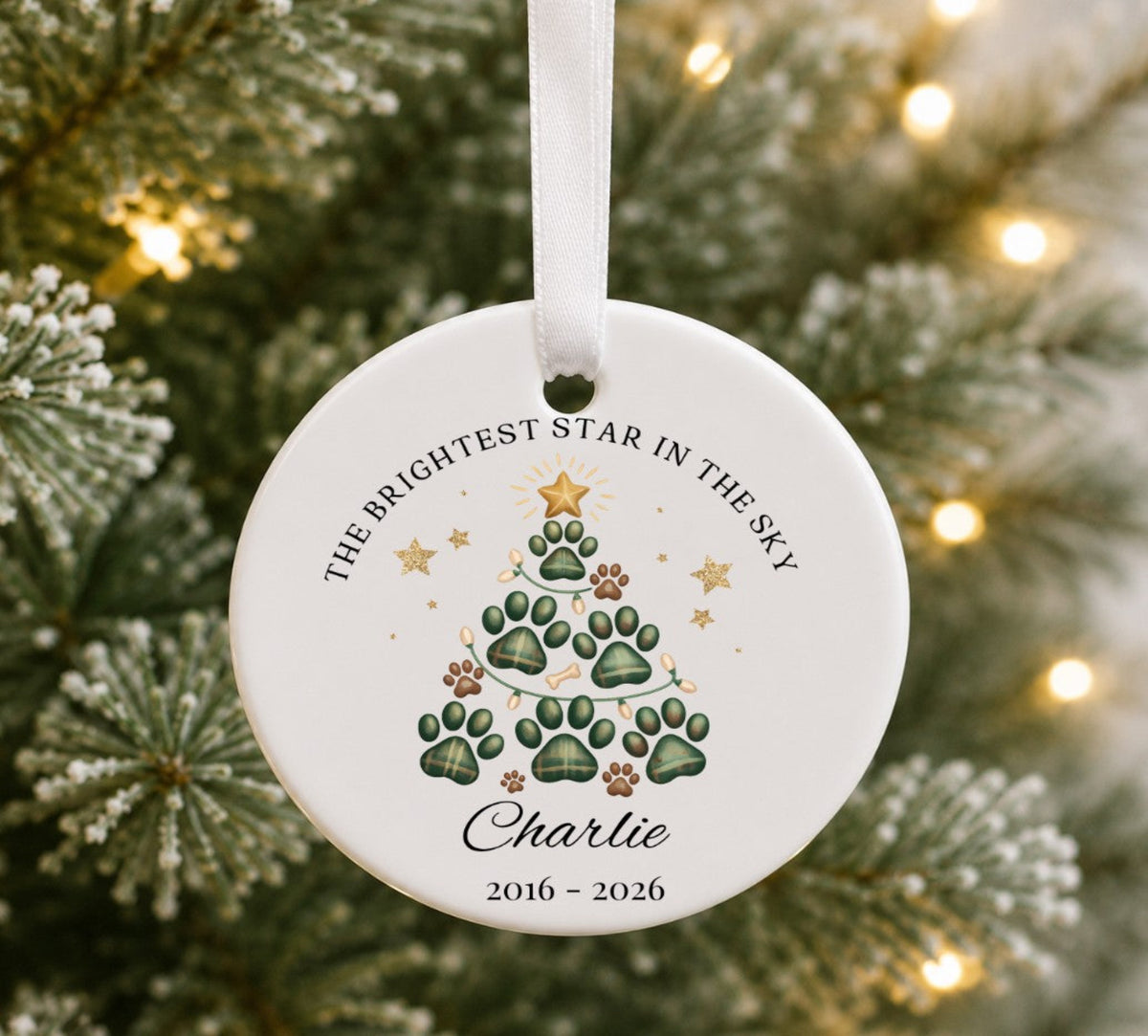 A personalised round white ceramic Christmas ornament hanging from a white ribbon, featuring a paw-print tree design with green paw prints, gold stars, and the text 'THE BRIGHTEST STAR IN THE SKY' and 'Charlie 2016 - 2026'. 