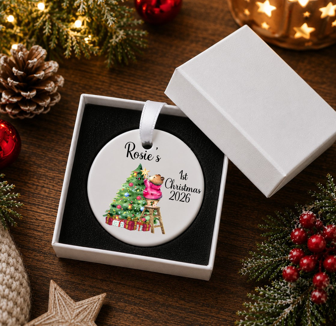 A round white ceramic Christmas ornament in a white gift box, printed with the name 'Rosie's', '1st Christmas 2026', and an illustration of a teddy bear decorating a Christmas tree.