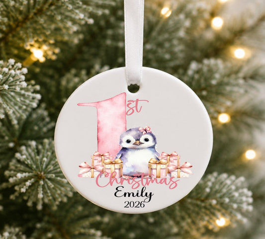 A personalised round ceramic Christmas ornament hanging on a tree. The white ornament features a watercolor illustration of a penguin with a pink bow, surrounded by pink gift boxes, a pink number 1, and the text '1st Christmas Emily 2026' in pink and black script.