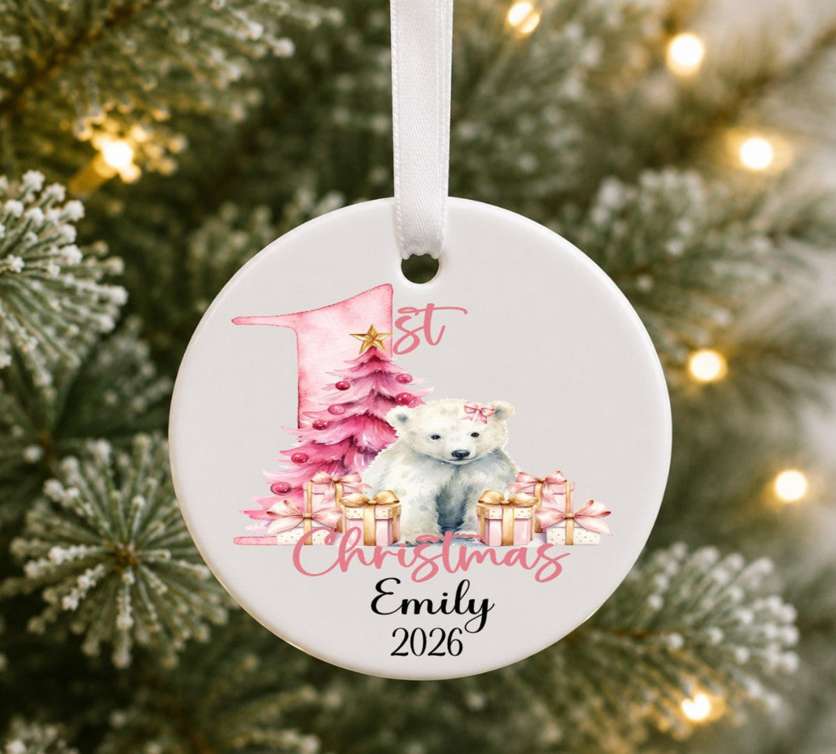 A personalised round white Christmas ornament hanging on a blurred Christmas tree with warm lights. The ornament features a watercolor polar bear, pink tree, gift boxes, and the text '1st Christmas Emily 2026'.