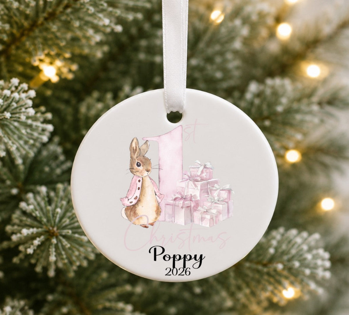 A round white ceramic Christmas ornament hanging from a white ribbon, featuring a watercolour illustration of Beatrix Potter's flopsy bunny Rabbit next to a pink number one and wrapped presents. The text 'Poppy 2026' is printed at the bottom.
