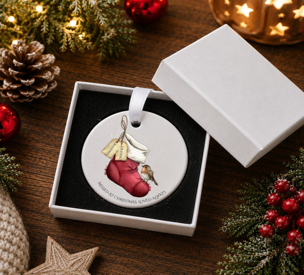 A round white ceramic Christmas ornament hanging from a white ribbon, featuring an illustration of a robin bird on a red stocking with two tags labelled 'GRANDMA and GRANDAD  'MISSED AT CHRISTMAS LOVED ALWAYS'. Presented in a white gift box.