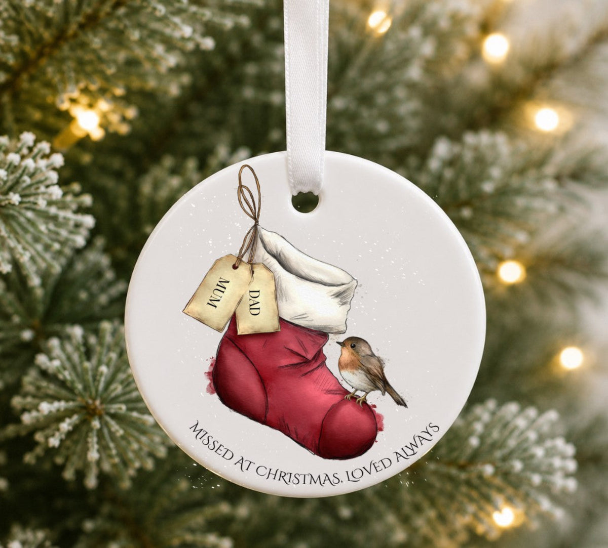 A round white Christmas ornament hanging from a white ribbon, featuring an illustration of a red stocking with a white trim, two hanging tags labelled 'DAD' and 'MUM', a robin bird, and the text 'MISSED AT CHRISTMAS, LOVED ALWAYS'. The ornament is photographed against a blurred Christmas tree background with warm lights.