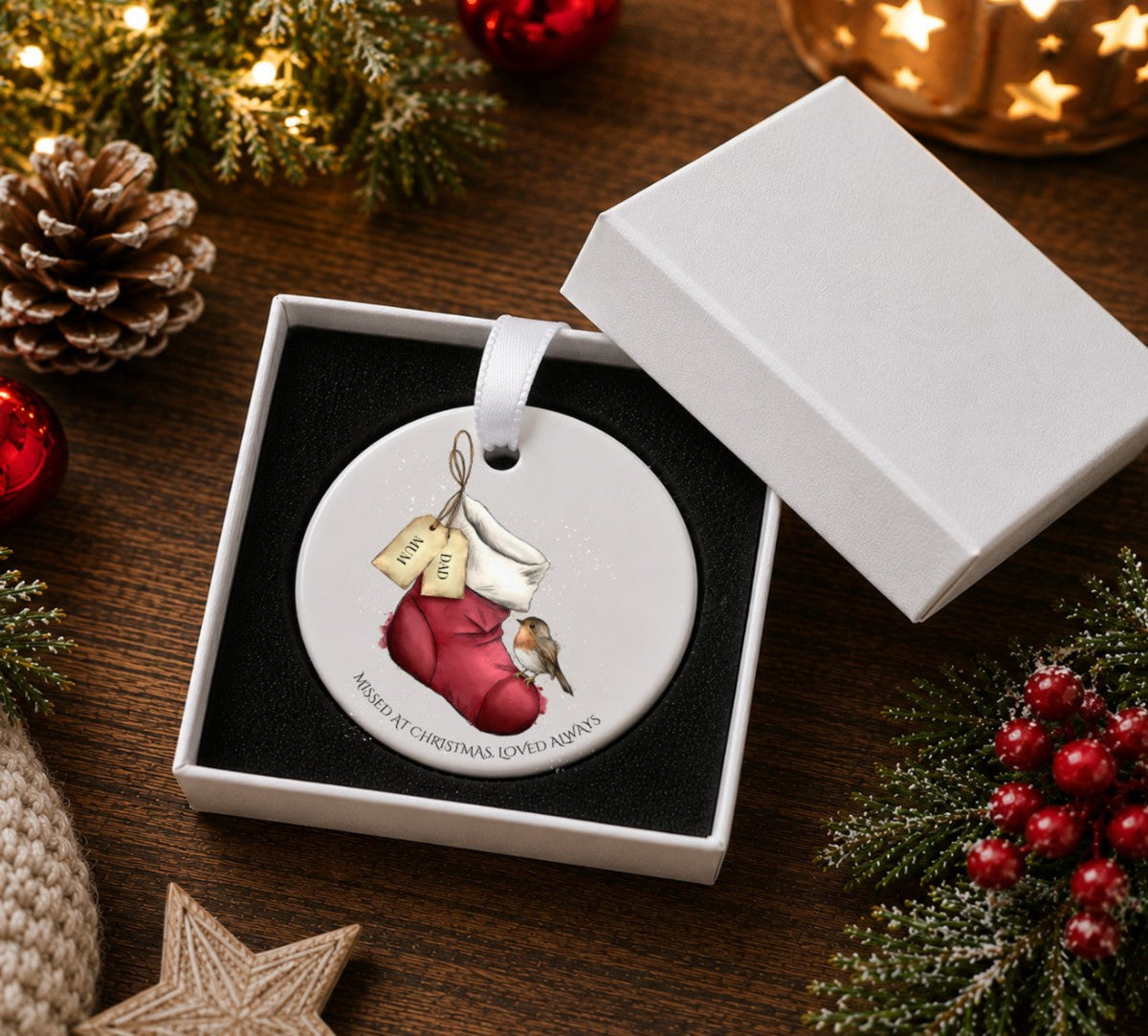A round white ceramic Christmas ornament hanging from a white ribbon, featuring an illustration of a robin bird on a red stocking with two tags labelled 'GRANDMA and GRANDAD  'MISSED AT CHRISTMAS LOVED ALWAYS'. Presented in a white gift box.