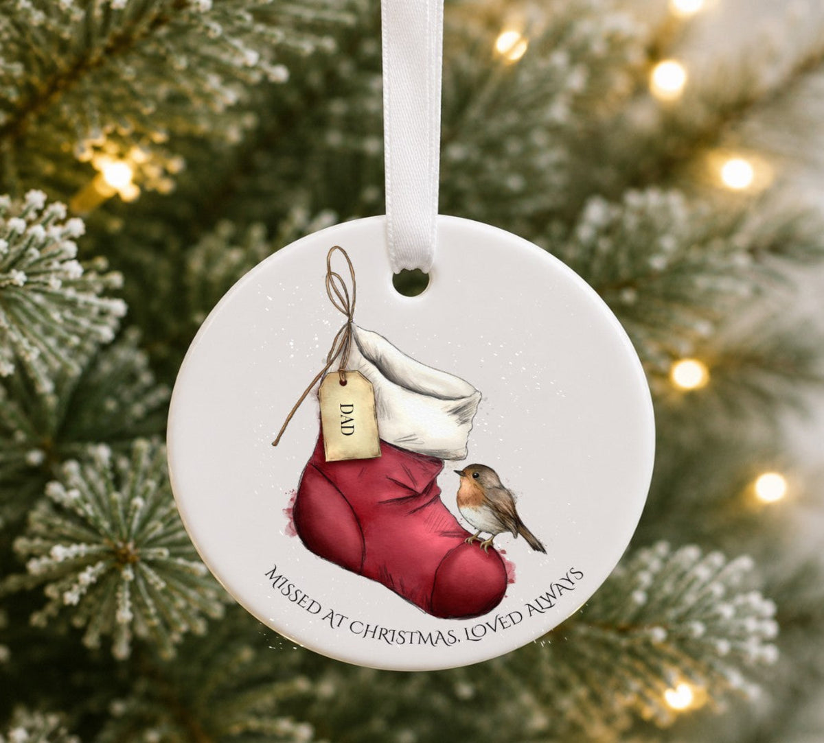 A round ceramic Christmas ornament. The ornament features an illustration of a  red Christmas stocking holding a small robin, with a hanging tag and the text 'MISSED AT CHRISTMAS, LOVED ALWAYS around the bottom edge.