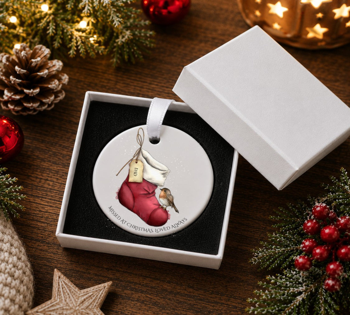 A round ceramic Christmas ornament displayed in an open white gift box. The ornament features an illustration of a  red Christmas stocking holding a small robin, with a hanging tag and the text 'MISSED AT CHRISTMAS, LOVED ALWAYS around the bottom edge.