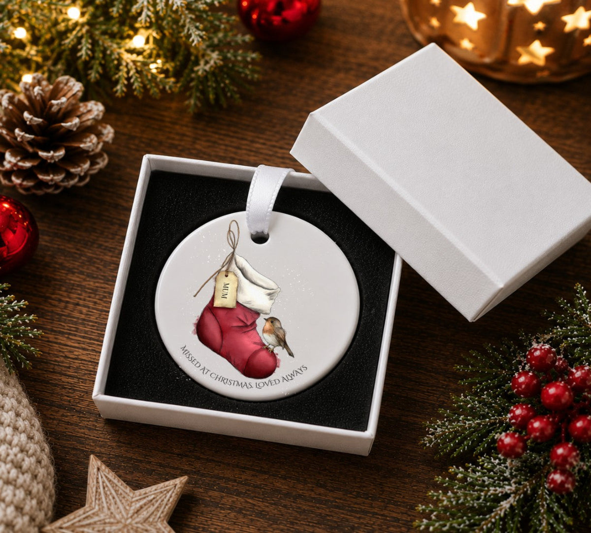 A round ceramic Christmas ornament displayed in an open white gift box. The ornament features an illustration of a  red Christmas stocking holding a small robin, with a hanging tag and the text 'MISSED AT CHRISTMAS, LOVED ALWAYS around the bottom edge.
