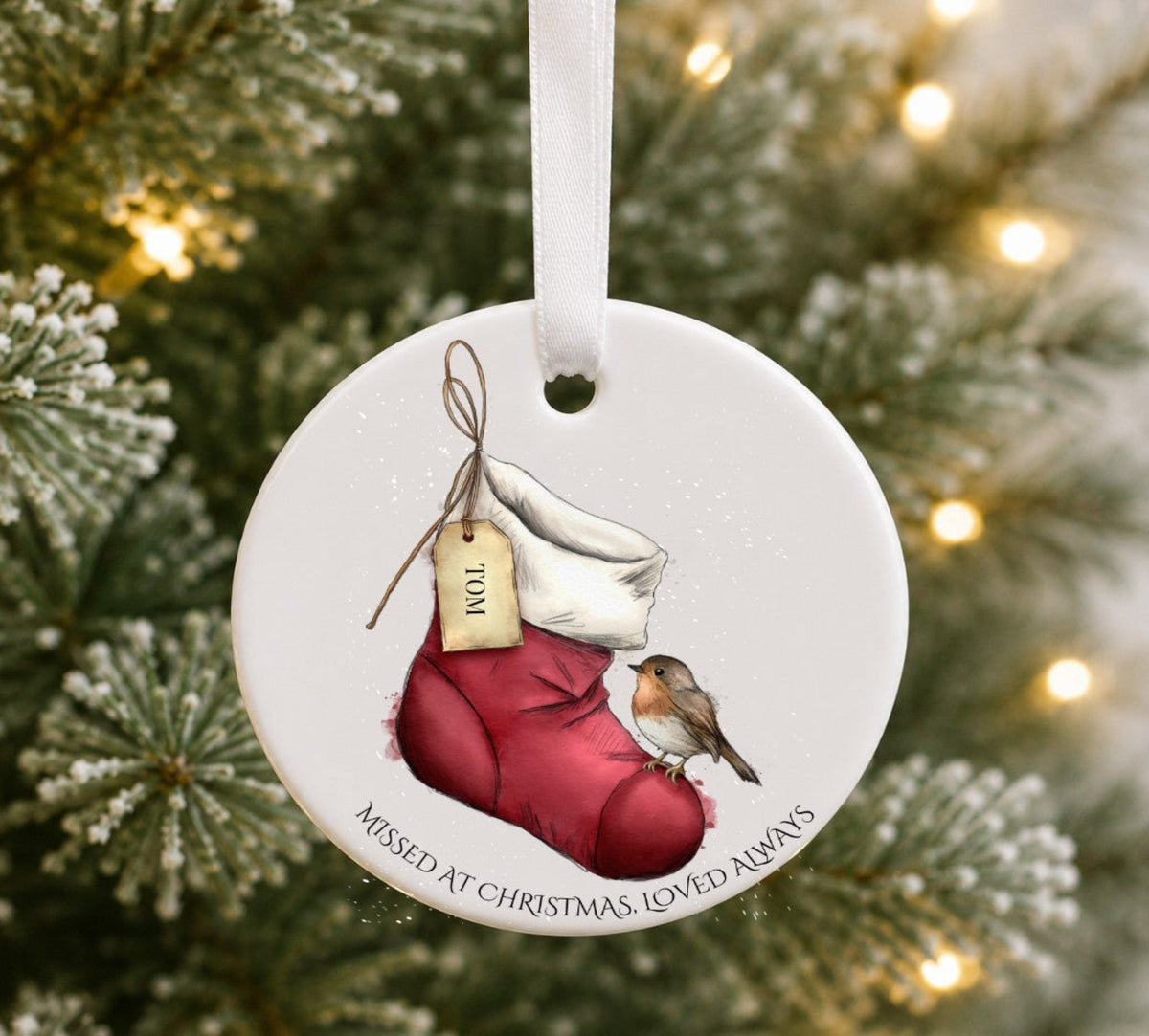 A round ceramic Christmas ornament . The ornament features an illustration of a  red Christmas stocking holding a small robin, with a hanging tag and the text 'MISSED AT CHRISTMAS, LOVED ALWAYS around the bottom edge.