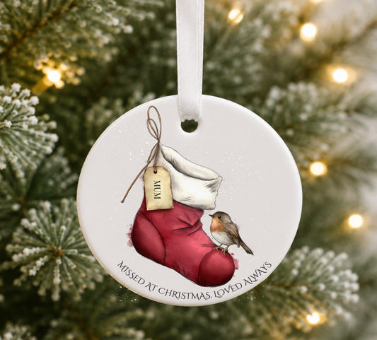 A round white ceramic Christmas ornament hanging from a white ribbon, featuring an illustration of a red stocking , a small robin bird, and the text 'MISSED AT CHRISTMAS LOVED ALWAYS'. The ornament is photographed against a blurred Christmas tree background with warm string lights.