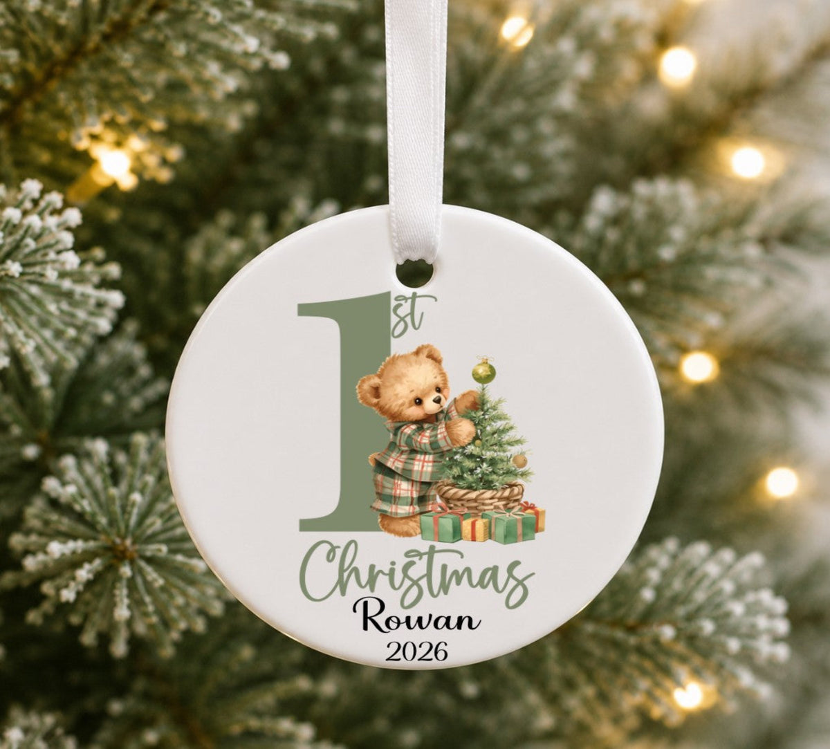 A personalised round ceramic Christmas ornament hanging on a blurred Christmas tree with warm lights. The white ornament features a printed illustration of a teddy bear pyjamas next to a small Christmas tree and presents, with the text '1st Christmas Rowan 2026' and a large number 1.
