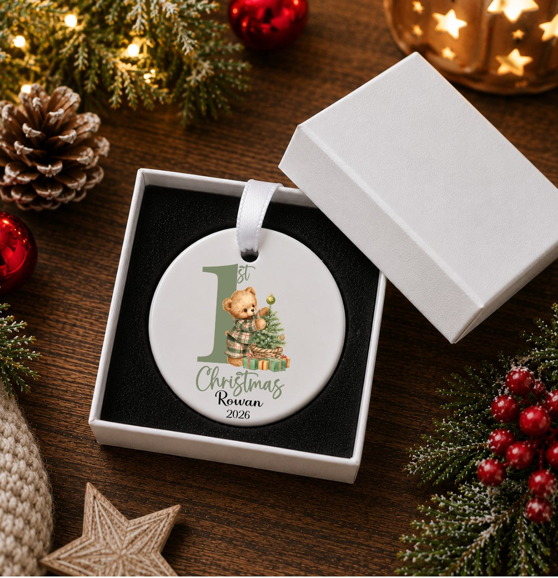 A round white Christmas ornament displayed in an open white gift box on a wooden surface, surrounded by holiday decorations including pine branches, red berries, a pinecone, and a lit star candle. The ornament features a teddy bear illustration and the text '1st Christmas Rowan 2026'.