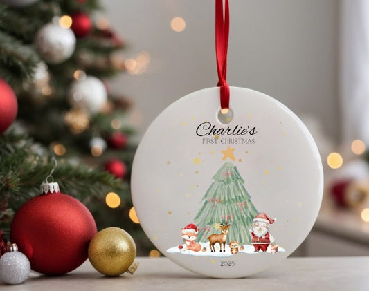 Personalised Christmas ornament with 'Charlie's First Christmas' text and festive woodland scene , hanging in front of a decorated tree.