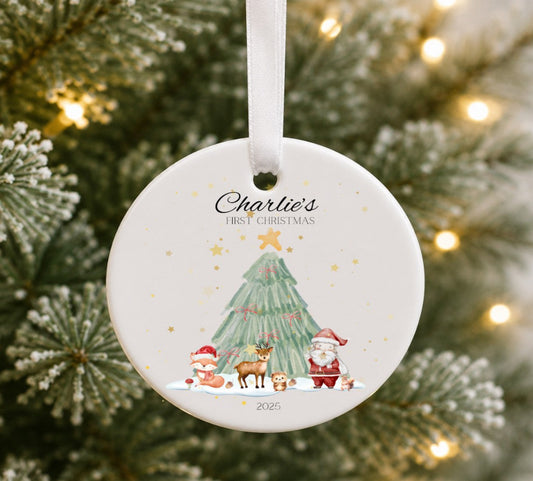 A personalised round Christmas ornament hanging from a white ribbon, featuring a watercolour illustration of a Christmas tree, Santa Claus, a fox, and a reindeer, with the text 'Charlie's First Christmas 2026'. The ornament is photographed in front of a blurred Christmas tree with warm string lights.